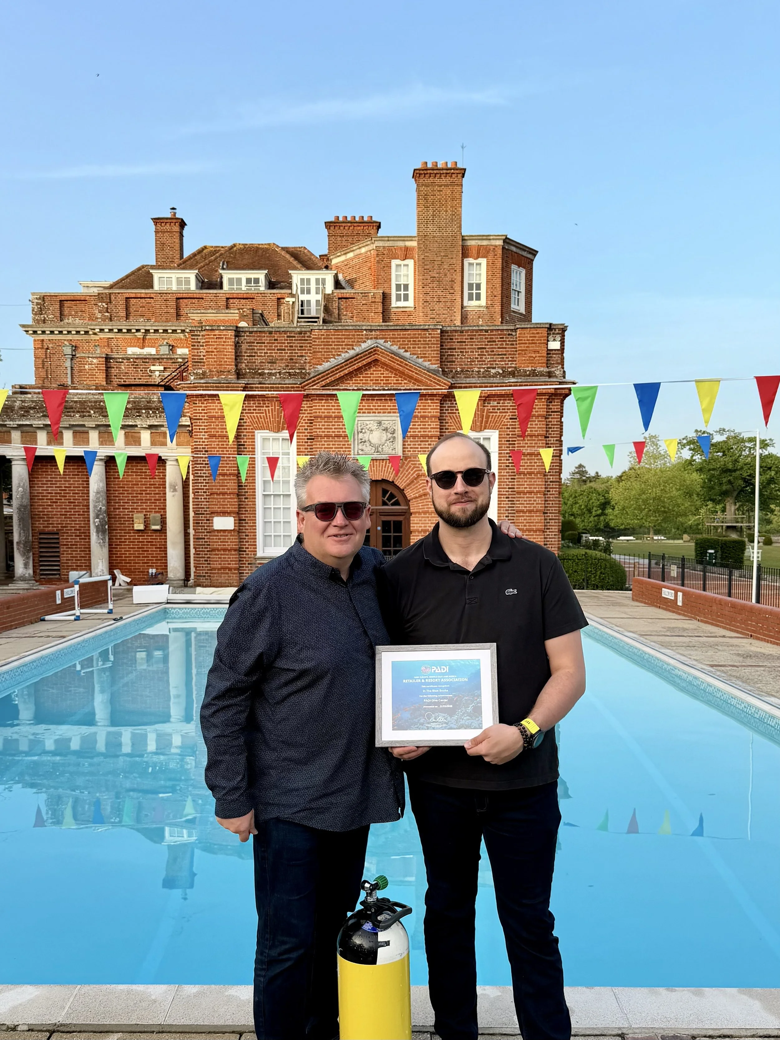 Two men standing in front of a swimming pool, holding a framed certificate. One is wearing sunglasses and a black Lacoste polo shirt, the other has gray hair and glasses. A yellow and black propane tank is on the ground in front of them. In the background is a red brick building with a blue sky and colorful bunting hanging overhead.