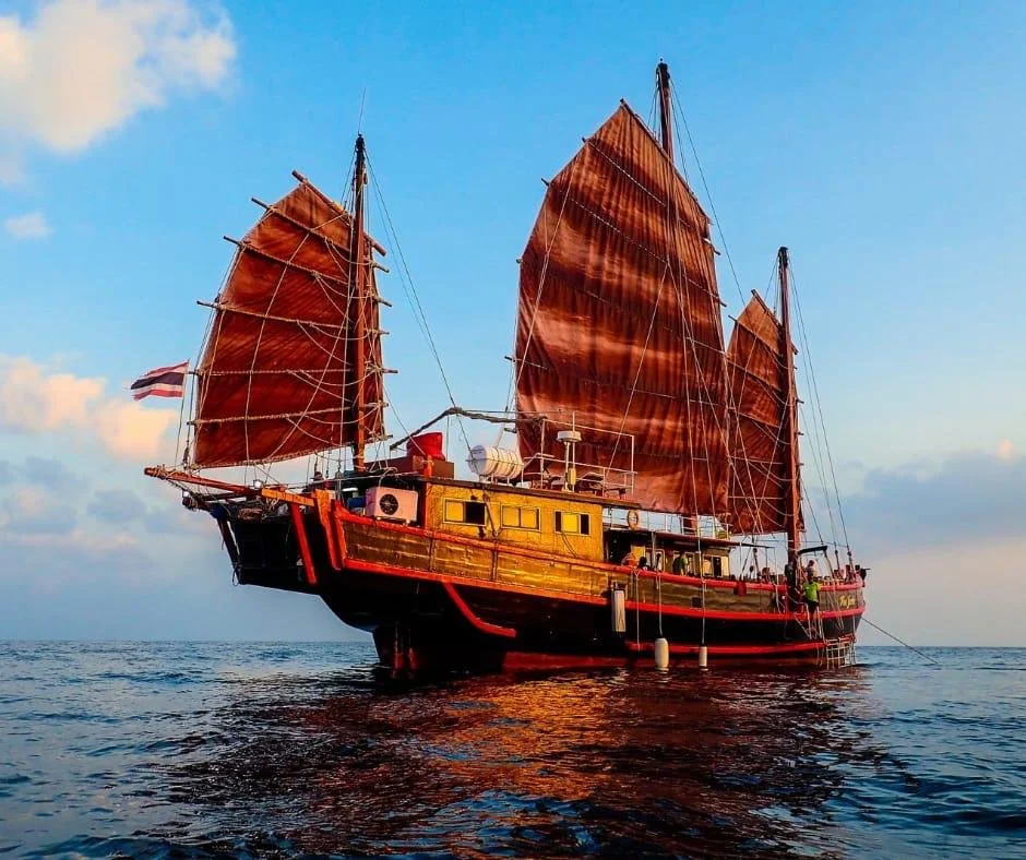 A traditional sailing ship with red sails floating on calm ocean water during sunset or sunrise.