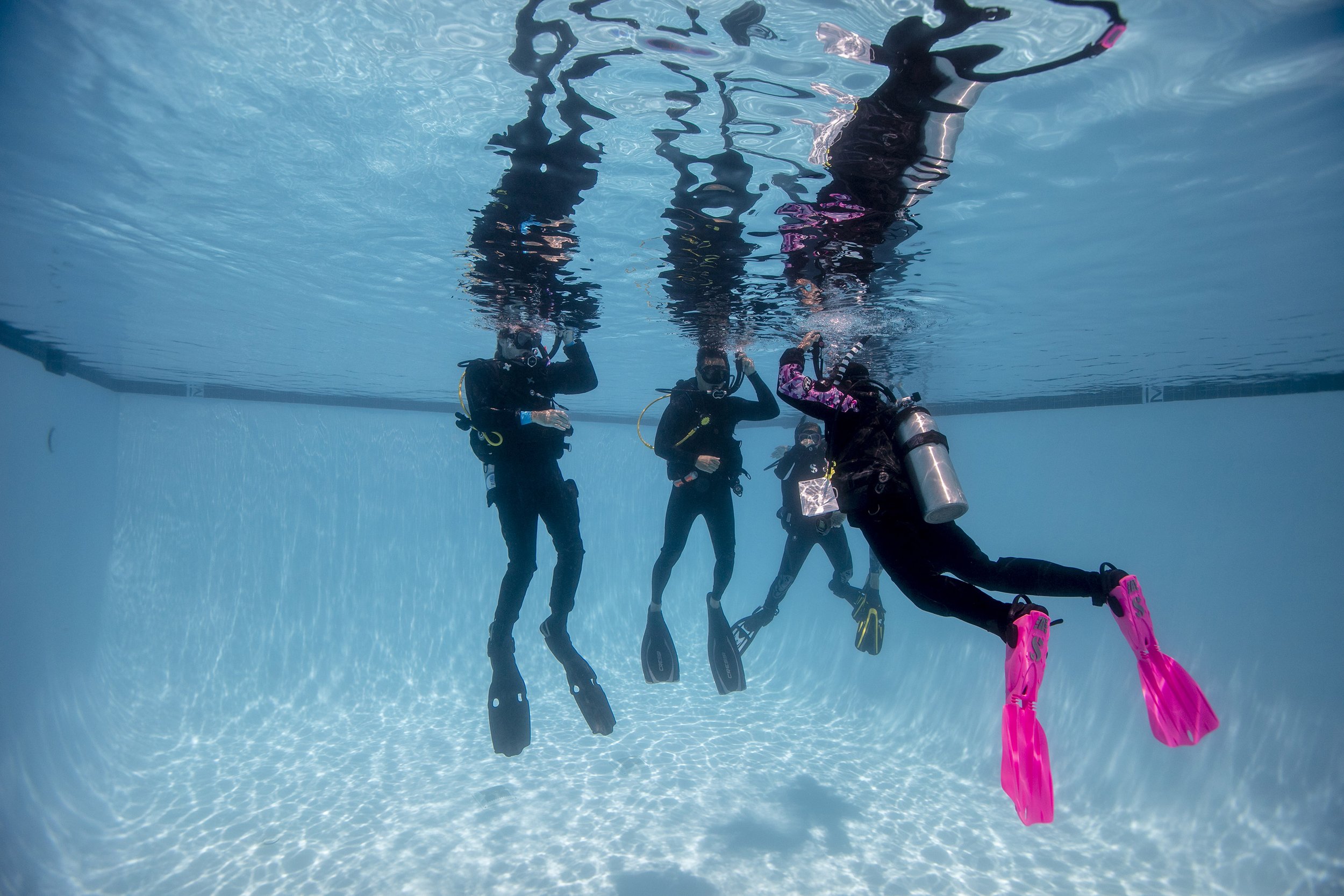 Group of four scuba divers underwater in a swimming pool, wearing diving suits and masks, with fins on their feet, posing with their hands forming various gestures.