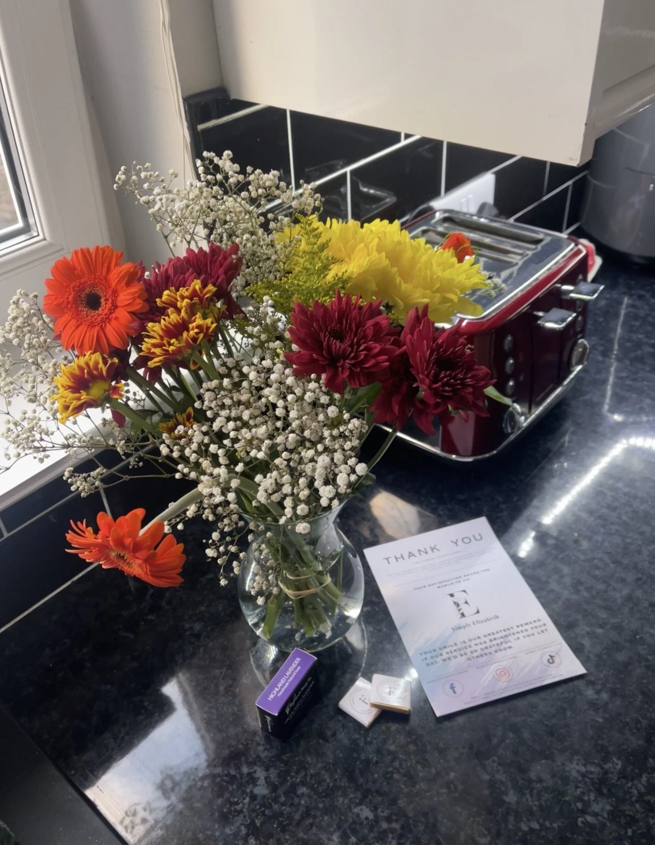 A kitchen countertop with a bouquet of mixed flowers in a glass vase, a thank you note, a small box of mints, and a toaster in the background.
