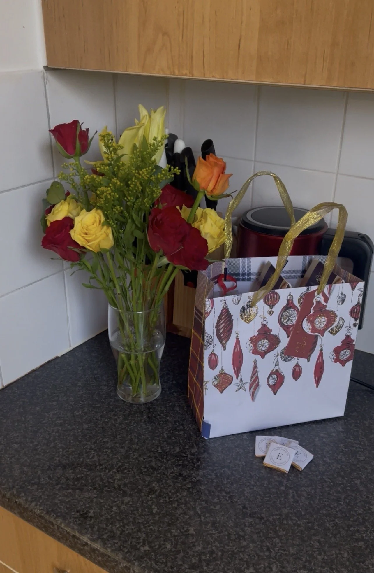 A kitchen countertop with a flower vase containing colorful roses, a holiday-themed gift bag, and some small packets.