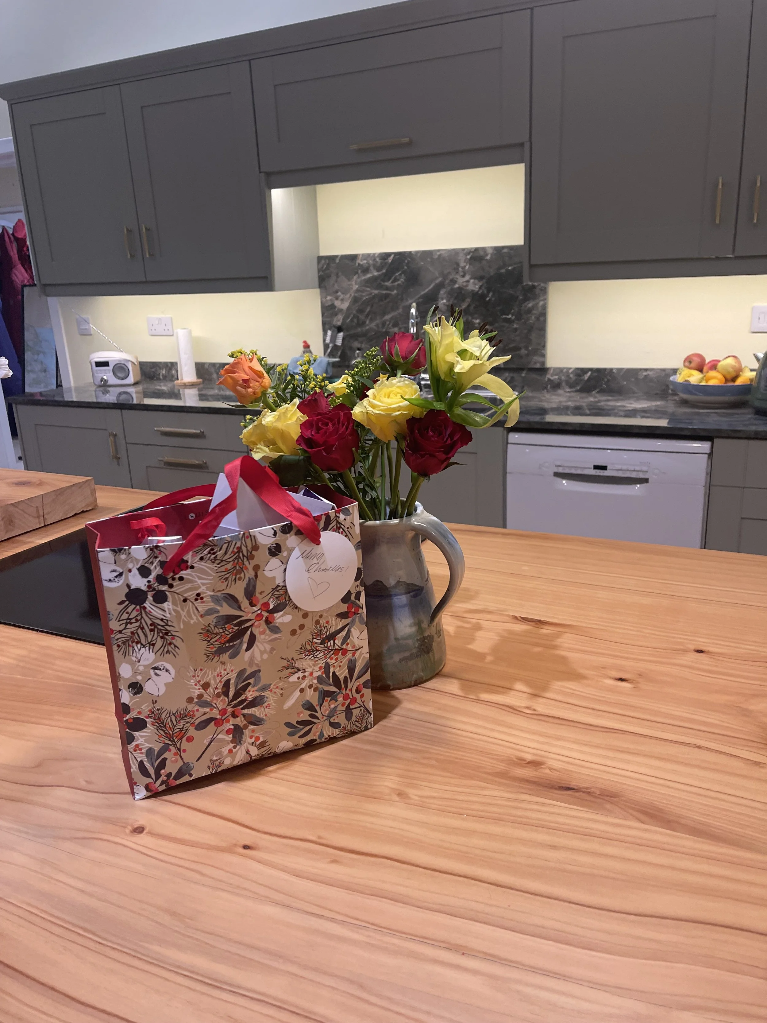 A gift bag and a vase with yellow, pink, and white flowers on a wooden kitchen island, with grey cabinets and marble backsplash in the background.