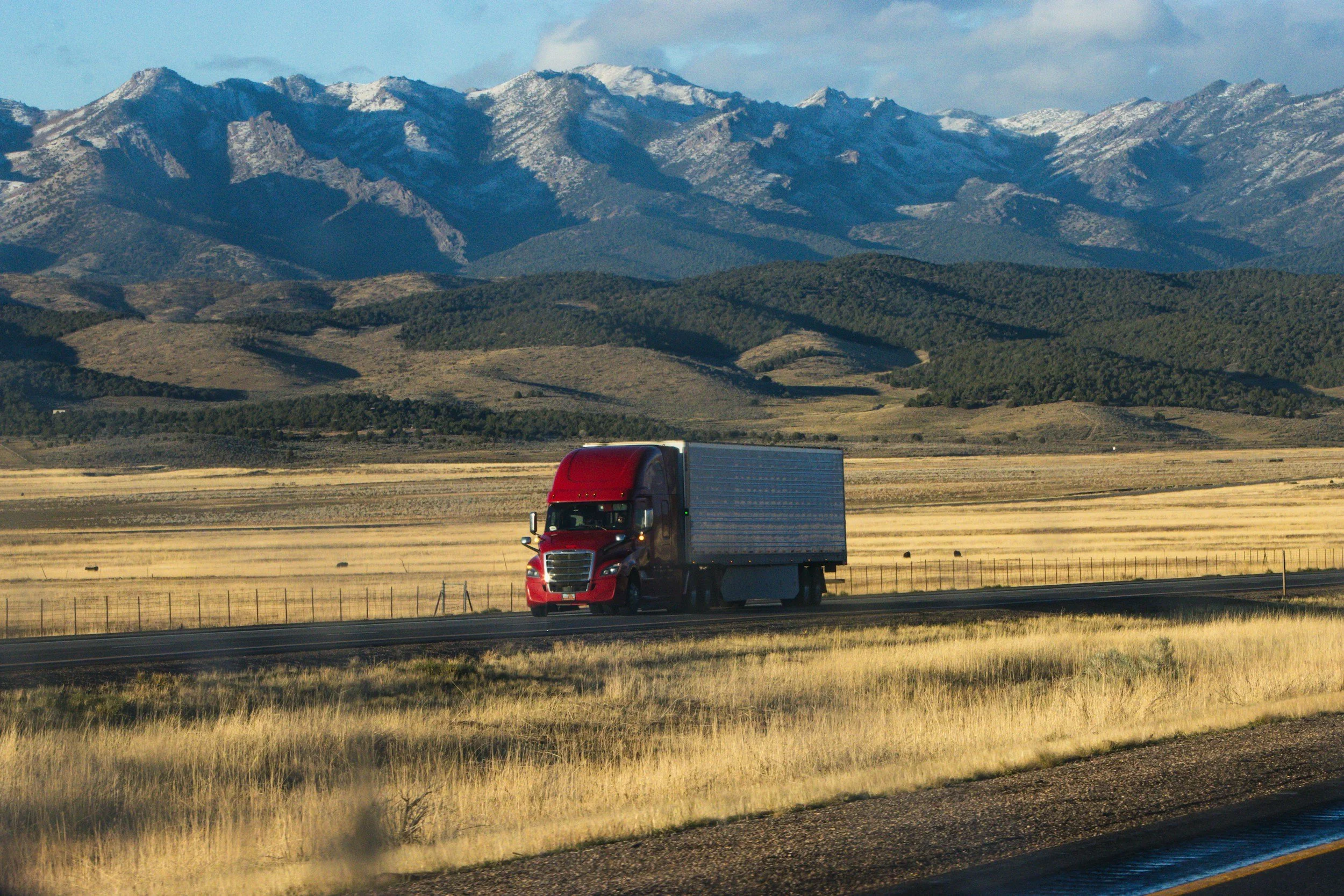 Red semi-truck driving on a highway through a landscape with yellow grass and mountains in the background.