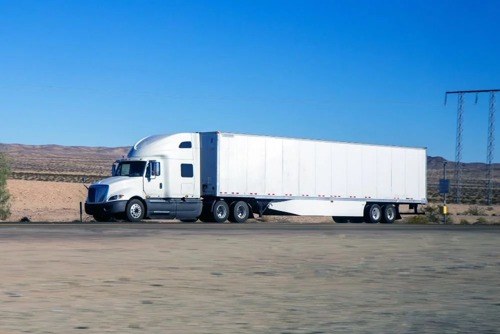 White semi-truck driving on a highway through a desert landscape with mountains and power lines in the distance.