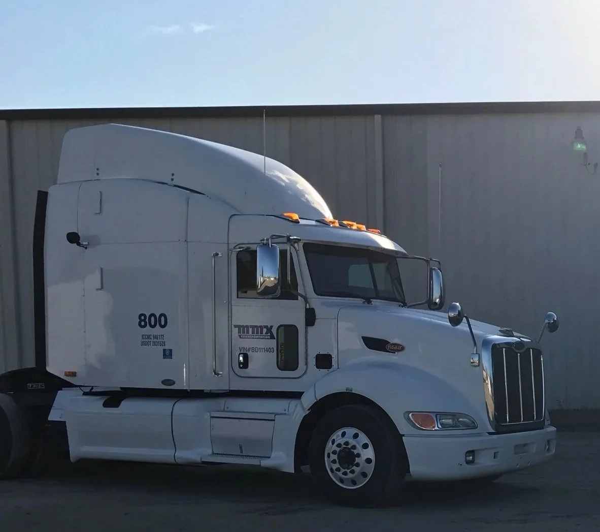 White semi-truck parked in front of a gray building with a green light on the wall, under a clear blue sky.
