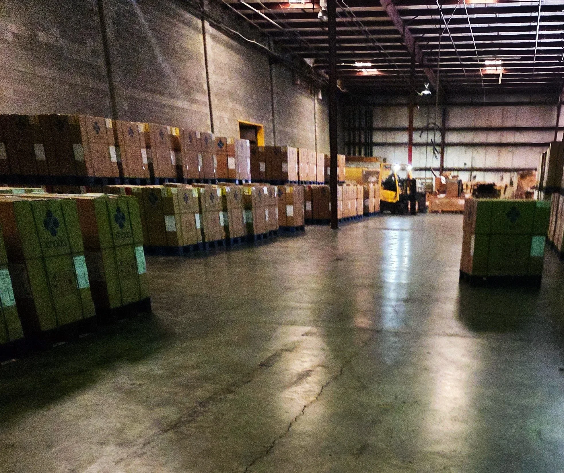 A warehouse with stacks of boxes on pallets, some loaded on trucks, with a concrete floor and high ceiling.