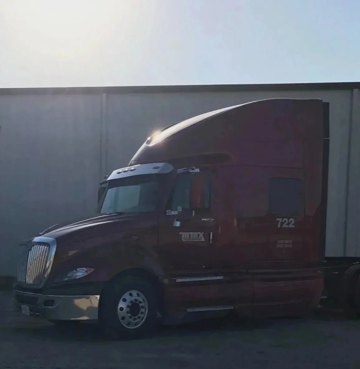 A dark red semi-truck parked on a street against a building wall, with sunlight reflecting off the front of the truck's cab.