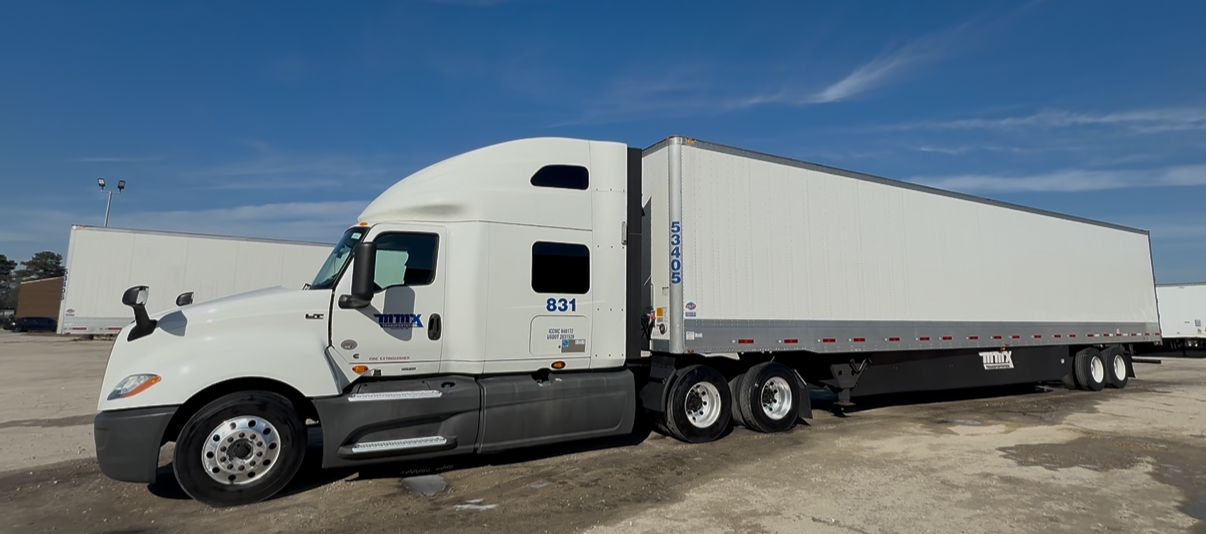 White semi-truck with a long trailer parked on a gravel lot under a blue sky.