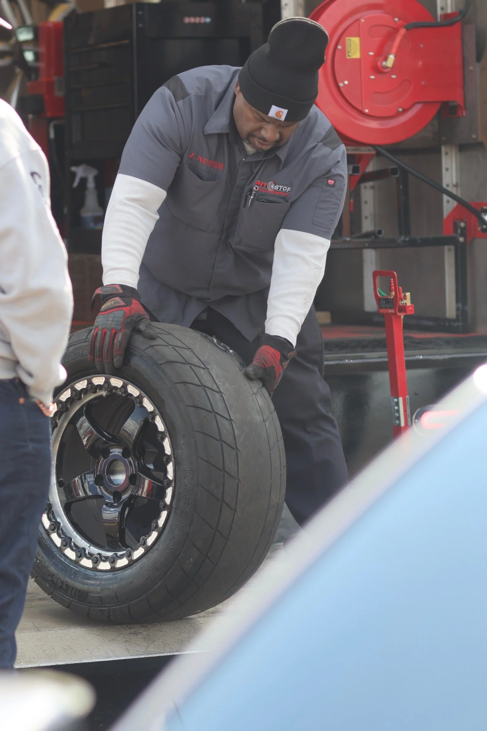 A man wearing a black beanie, gray Carhartt work shirt, and red gloves is changing a racing tire outdoors with a man in white and a red tire-changing machine visible nearby.