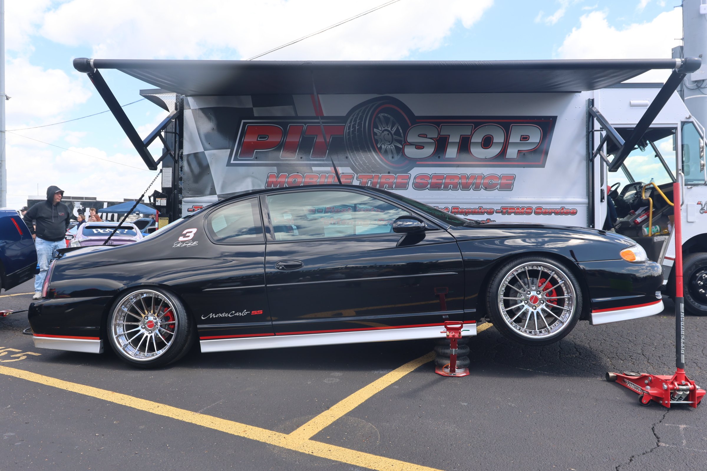 Black Chevrolet Monte Carlo SS race car elevated on jack stands in front of a pit stop truck at a racing event. The car has racing wheels, red brake calipers, and decals, with a background of people and other vehicles.