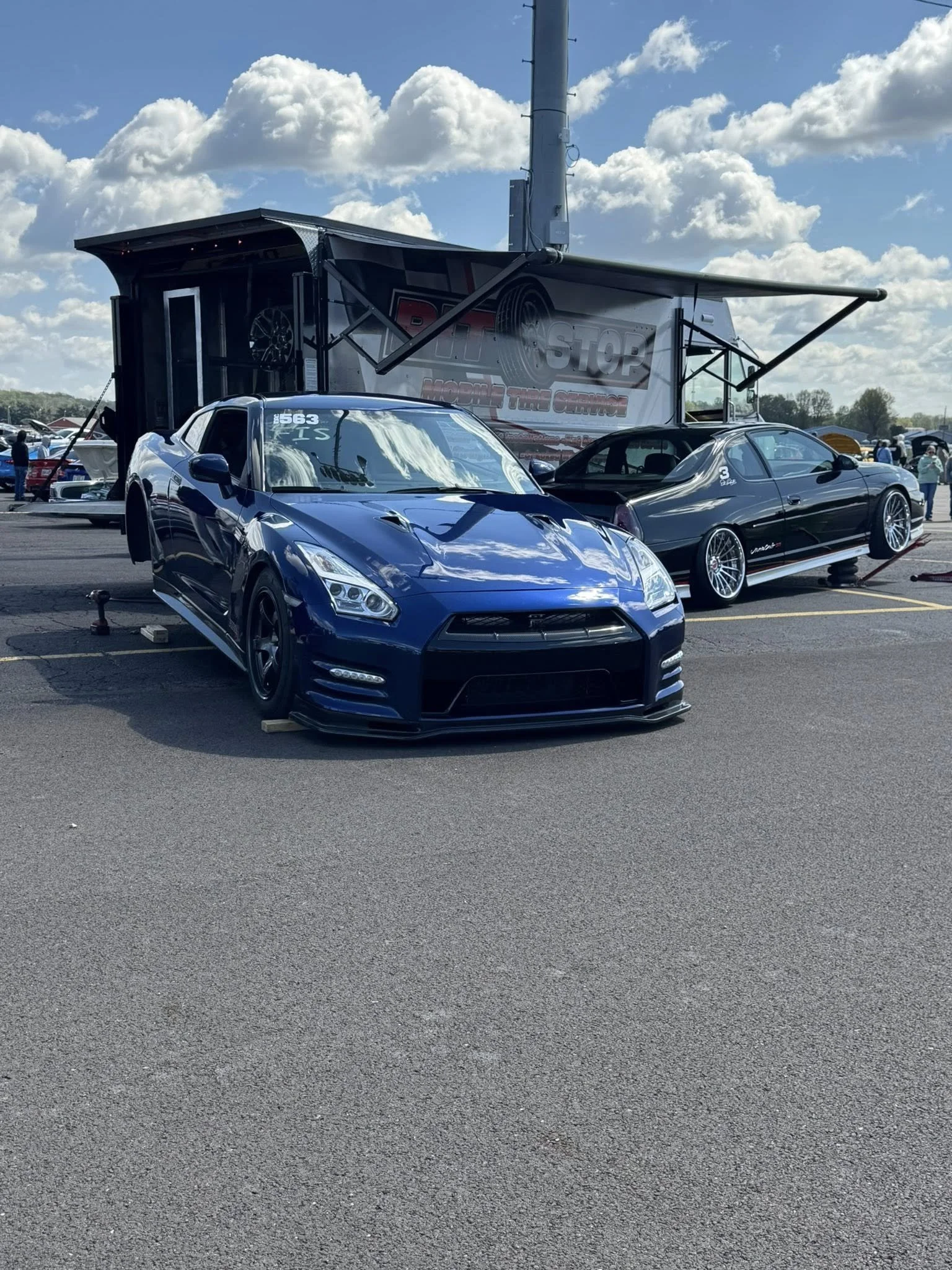 A blue sports car parked in a racing event with a black car beside it, both on a paved surface under a partly cloudy sky.