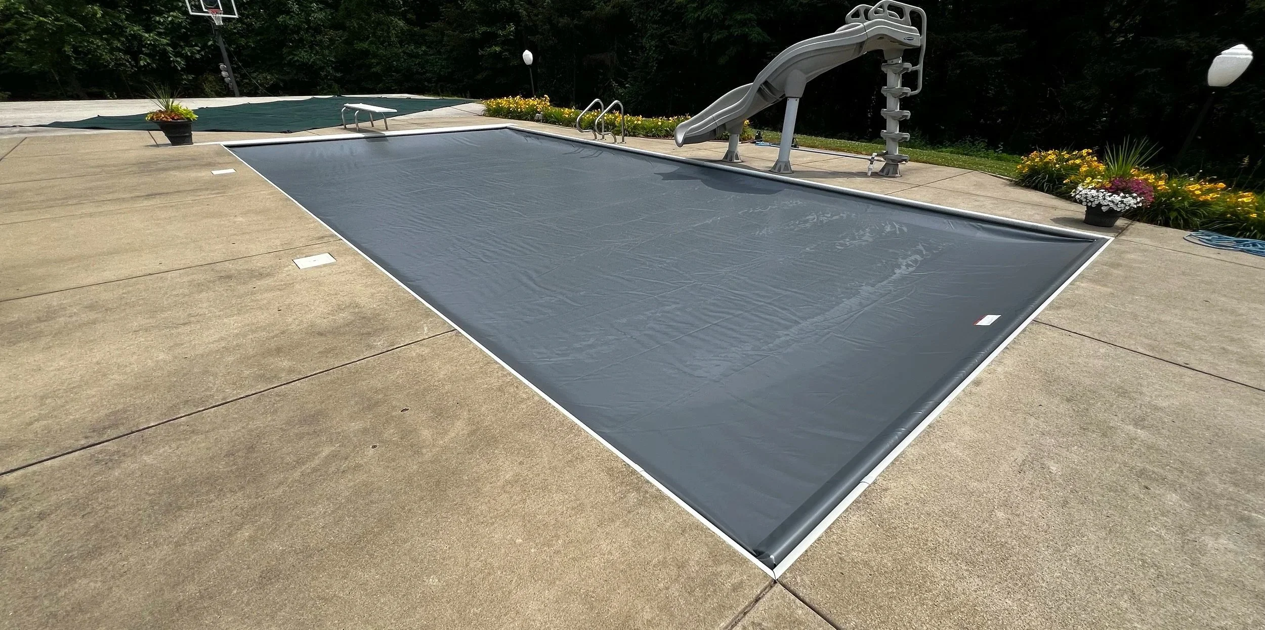 An empty backyard swimming pool covered with a gray safety cover, surrounded by a concrete deck, with a small water slide at one end, and flower pots with colorful flowers along the edges.