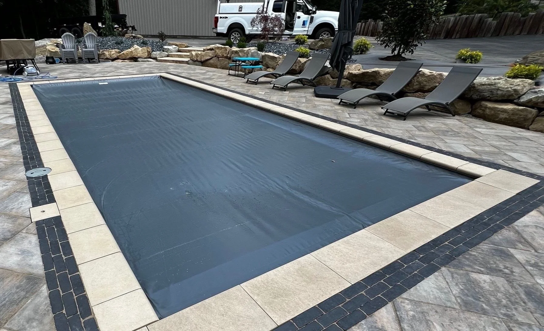 Covered rectangular backyard swimming pool with beige tiles and black accents, surrounded by lounge chairs, rocks, potted plants, a patio umbrella, and a parked white pickup truck in the background.