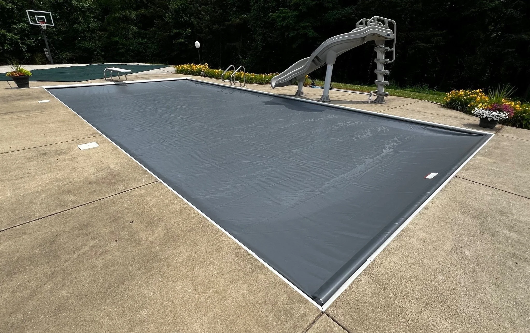 A backyard swimming pool with a cover, a water slide, and a basketball hoop in the background, surrounded by potted plants and flowers, and a wooded area behind.
