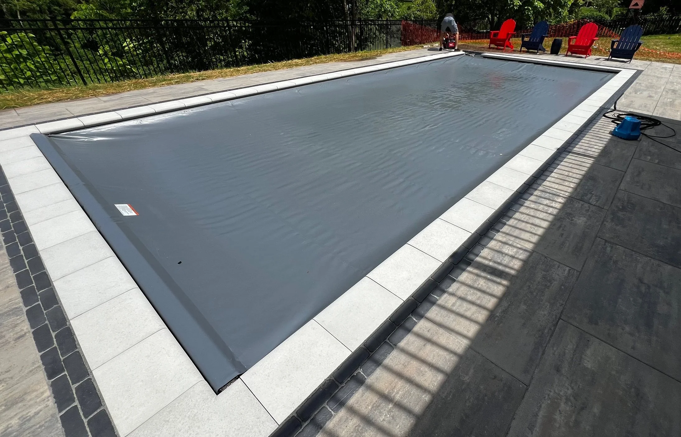 A backyard swimming pool covered with a gray safety cover, surrounded by a paved patio area with chairs at the far end.