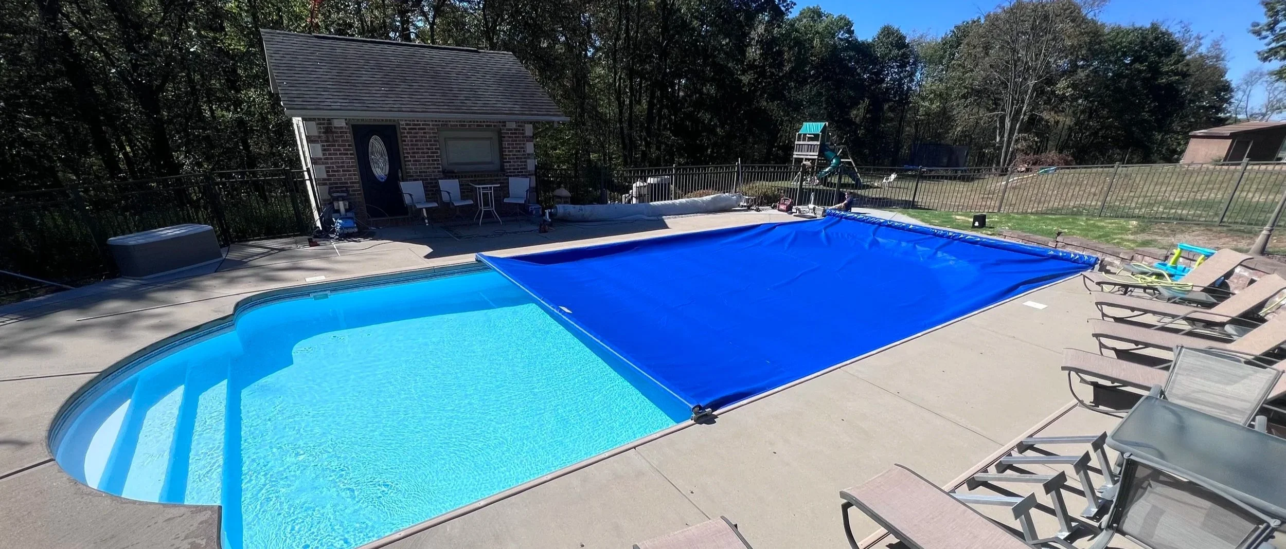 Empty residential swimming pool with a blue cover partially pulled over it, surrounded by a concrete deck with lounge chairs, and a fenced backyard with trees and a small shed.