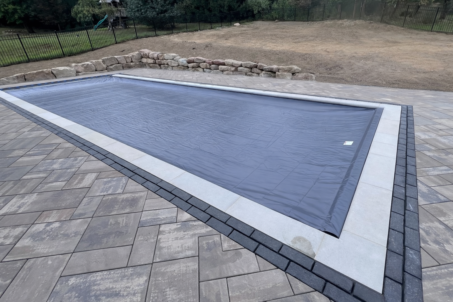 A swimming pool covered with a black automatic pool cover, surrounded by a paved stone patio and a stone border, in a backyard enclosed by a black fence.