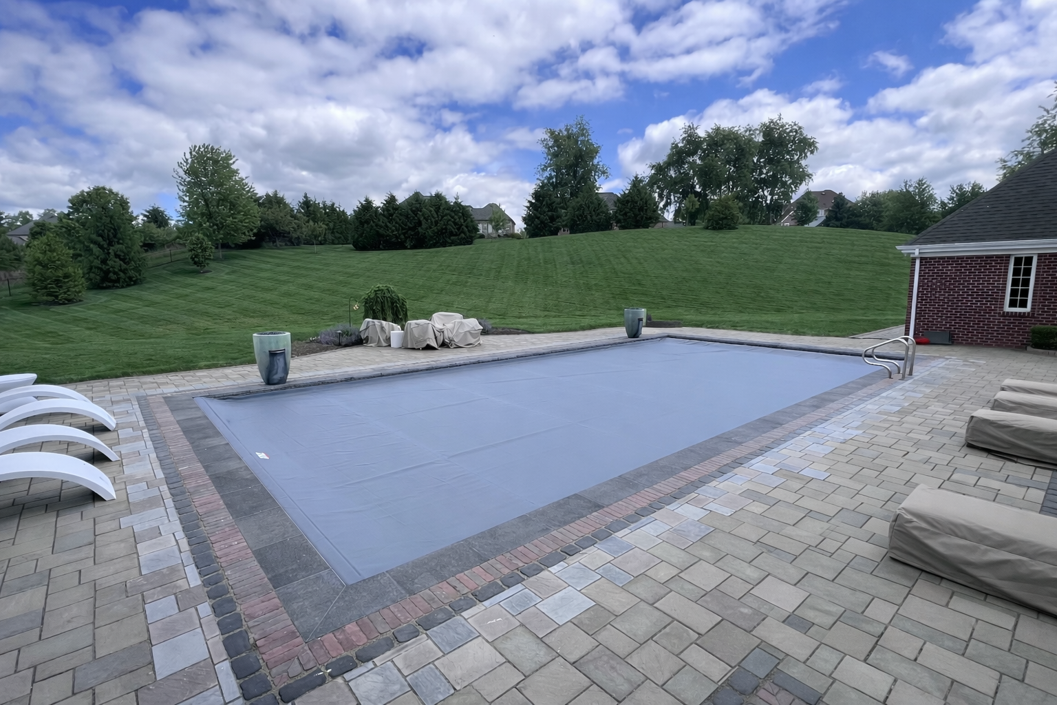 Backyard swimming pool covered with an automatic cover, surrounded by a patio with lounge chairs, against a backdrop of a grassy hill and a partly cloudy sky.