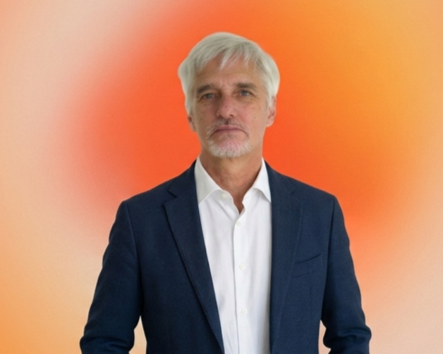A middle-aged man with gray hair and beard wearing a dark blazer and white shirt standing against a gradient orange background.