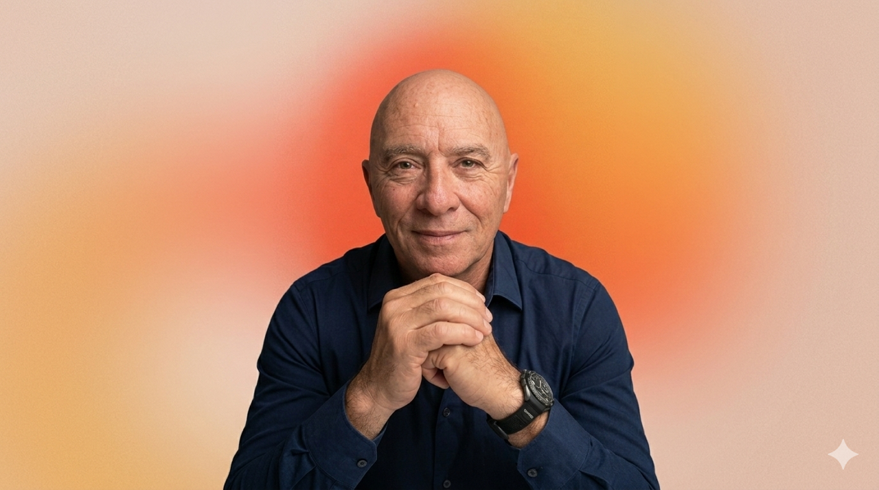 A bald man in a navy blue shirt, sitting with hands clasped, smiling slightly against a colorful gradient background.