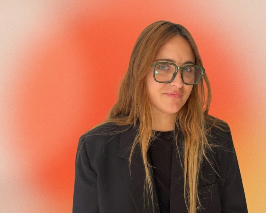 A woman with long red hair, glasses, and a black jacket, standing against a blurred orange and cream background.