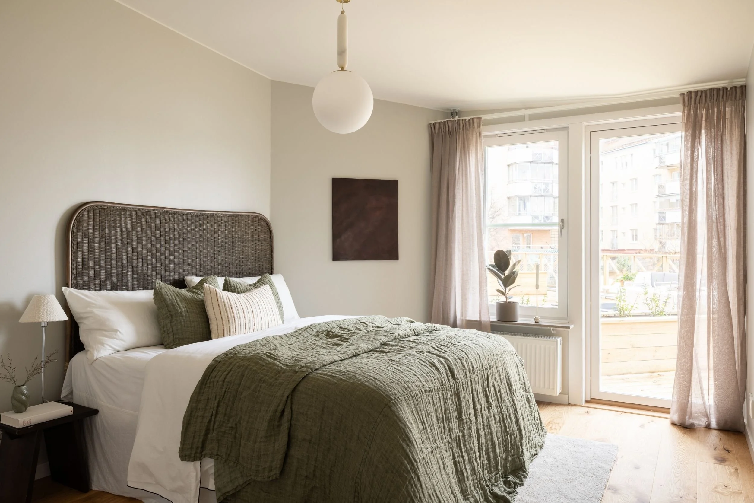 Sovrum med stor säng med vita täcken och kuddar, grönt överkast och prydnadskuddar, nattduksbord med lampa och böcker samt stora fönster med ljusa gardiner och växt på fönsterbrädan med utsikt mot byggnader