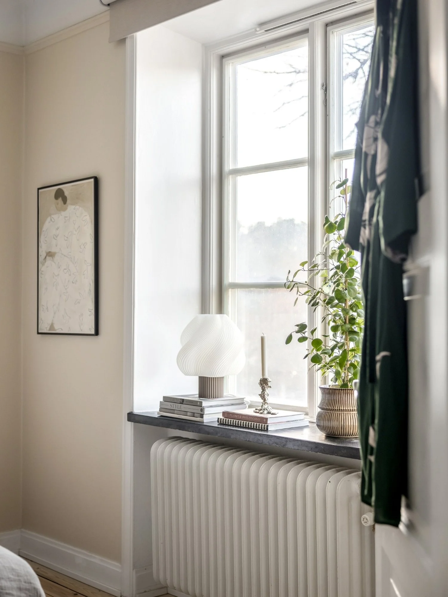 Fönster med stort ljusinsläpp i lägenhet stylat med modern lampa, krukväxt, böcker och ljusstake