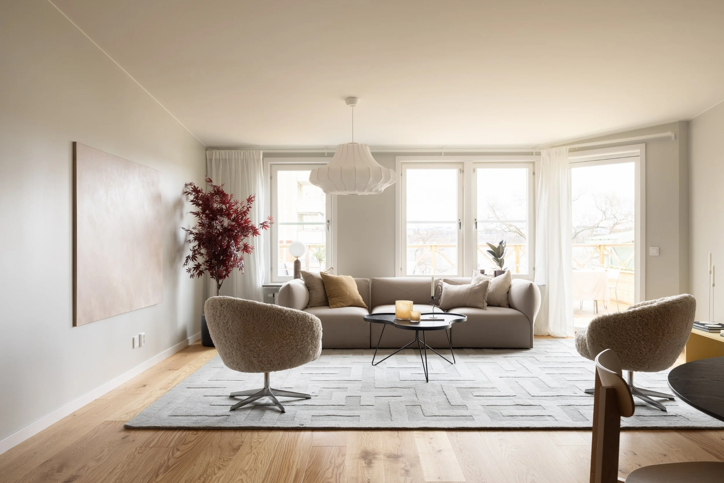 Ljust och luftigt vardagsrum med stora fönster, ljus soffa, två fåtöljer i fårskinn, stor matta och dekorativa detaljer