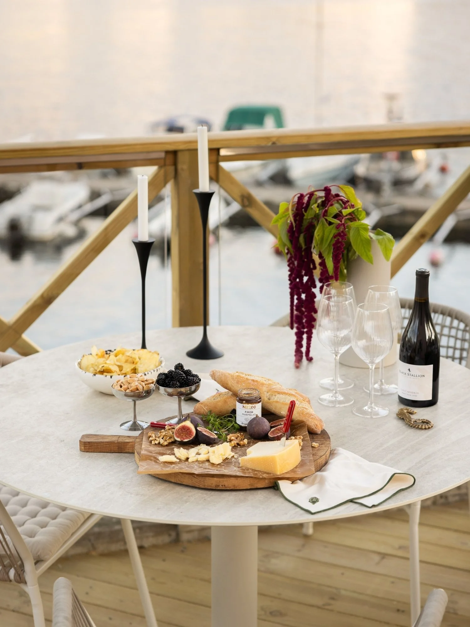 Matbord på uteplats med havsutsikt, stylat med ljusstakar, bukett i vas, vinflaska, fyra vinglas, snacks och charkbricka