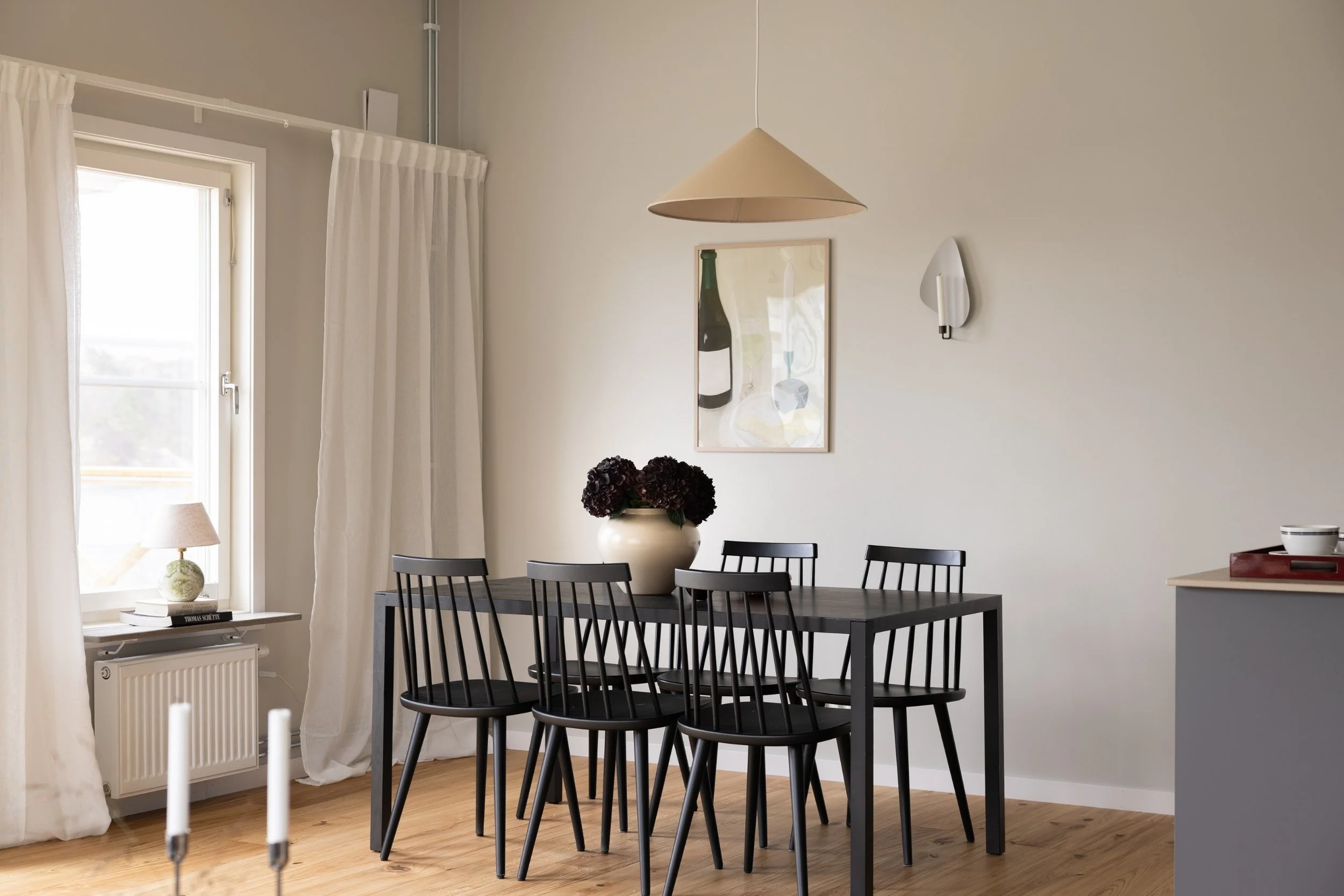Matplats med svart matbord, sex svarta pinnstolar, stor vas med mörka blommor, tavla och väggljusstake samt stort fönster med vita gardiner och lampa på böcker på fönsterbrädan