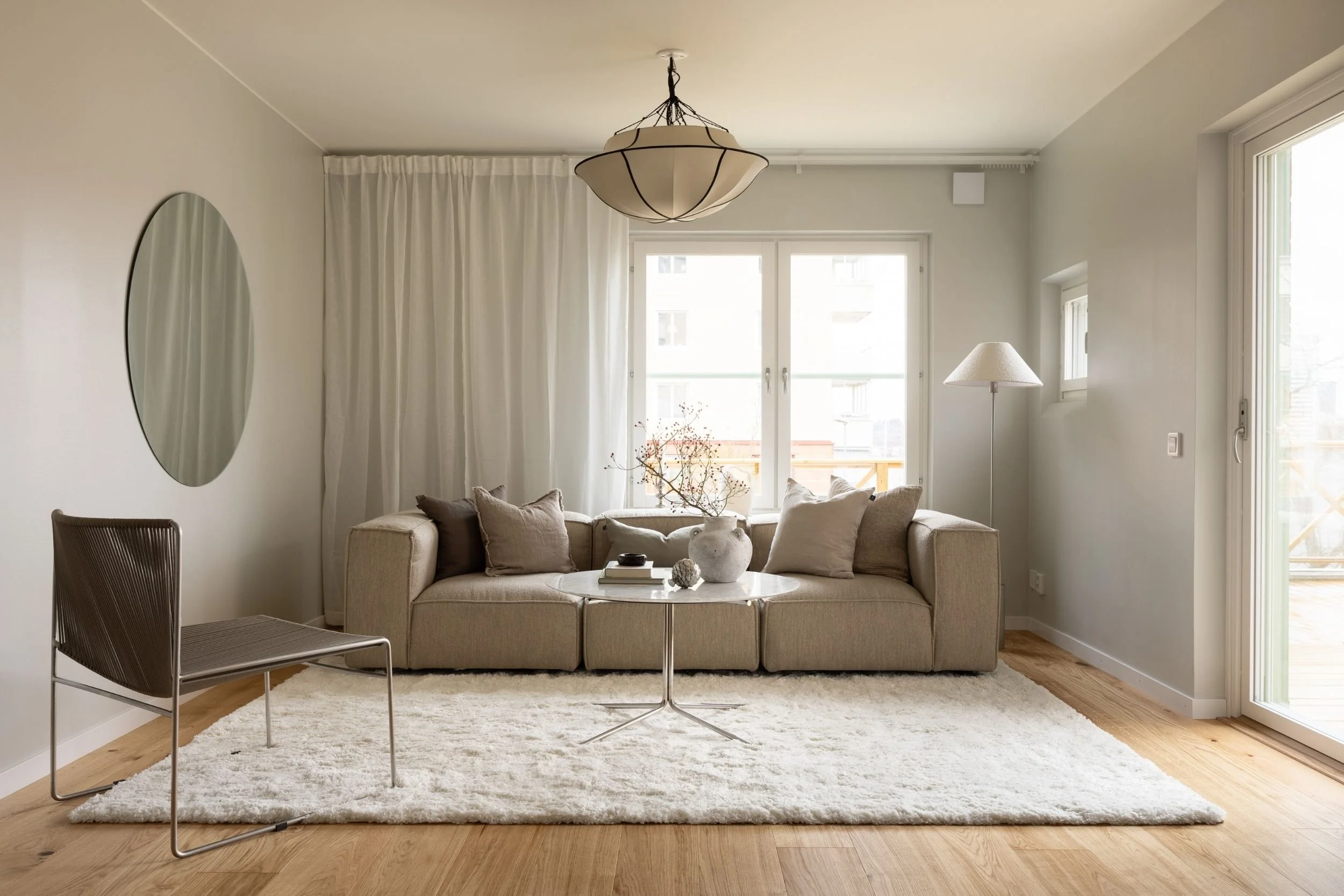 Skandinaviskt vardagsrum med ljus beige soffa, modern fåtölj, runt soffbord i marmor och stål med dekor, vit matta, taklampa och golvlampa, stor rund spegel samt fönster och dörr med utsikt mot uteplats