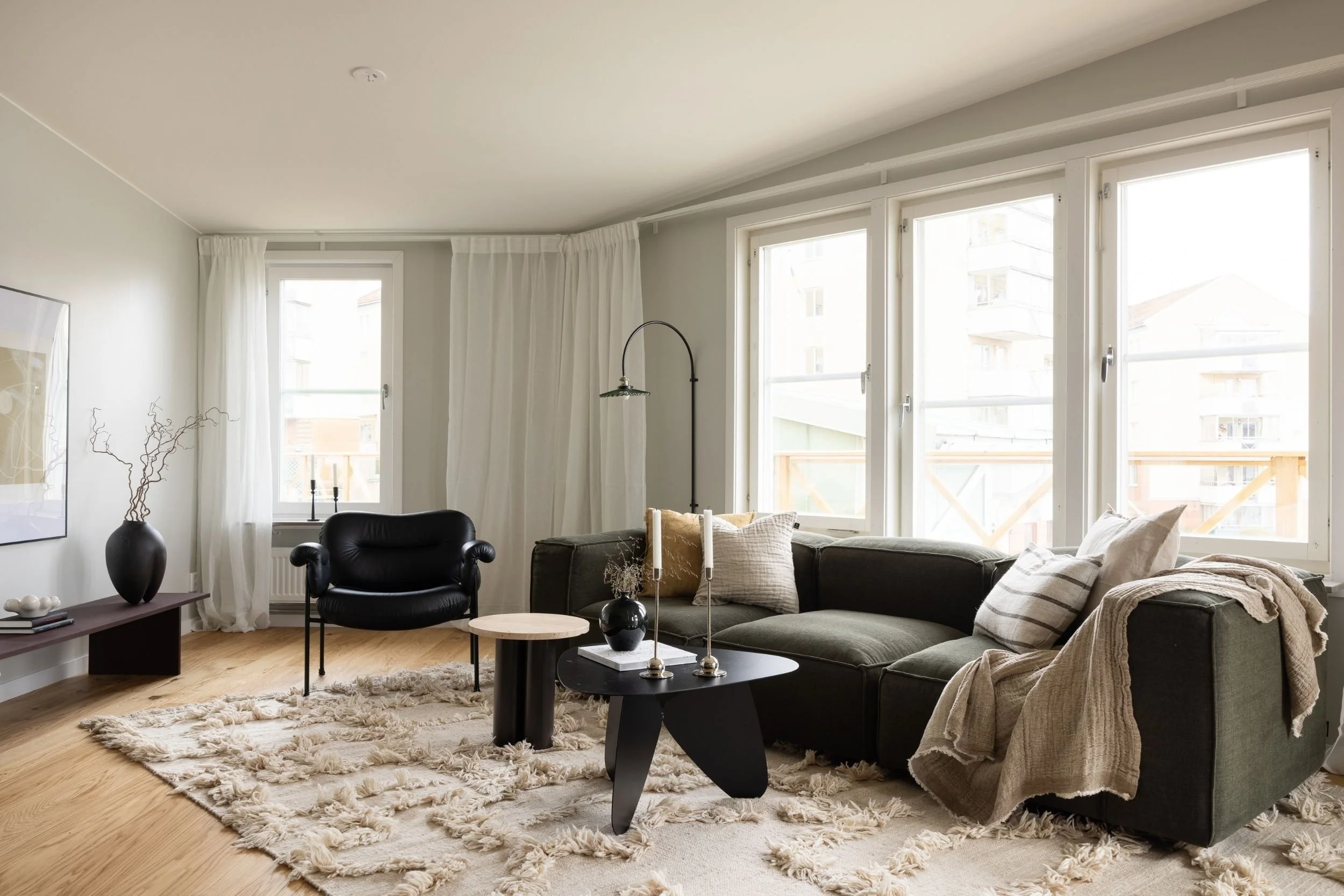 Vardagsrum med mörk låg soffa och fåtölj, två runda soffbord, stor vägglampa, vita gardiner, stora fönster, ljust trägolv, beige ullmatta, väggkonst samt svarta vaser och ljusstakar