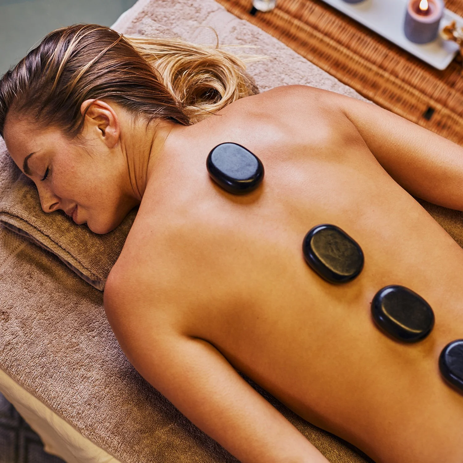 A woman lying face down on a massage table with four black hot stones placed along her back at Renewed Life Esthetics and Head Spa.