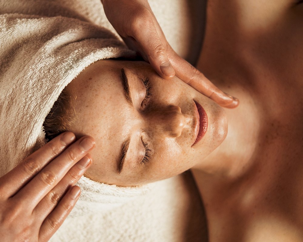 Close-up of a woman receiving a facial massage with a towel wrapped around her head at Renewed Life Esthetics and Head Spa.