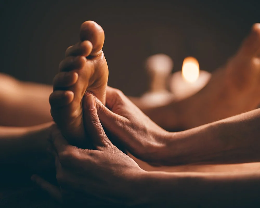 Close-up of a person receiving a massage, hand pressing on their foot in a dimly lit room at Renewed Life Esthetics and Head Spa.
