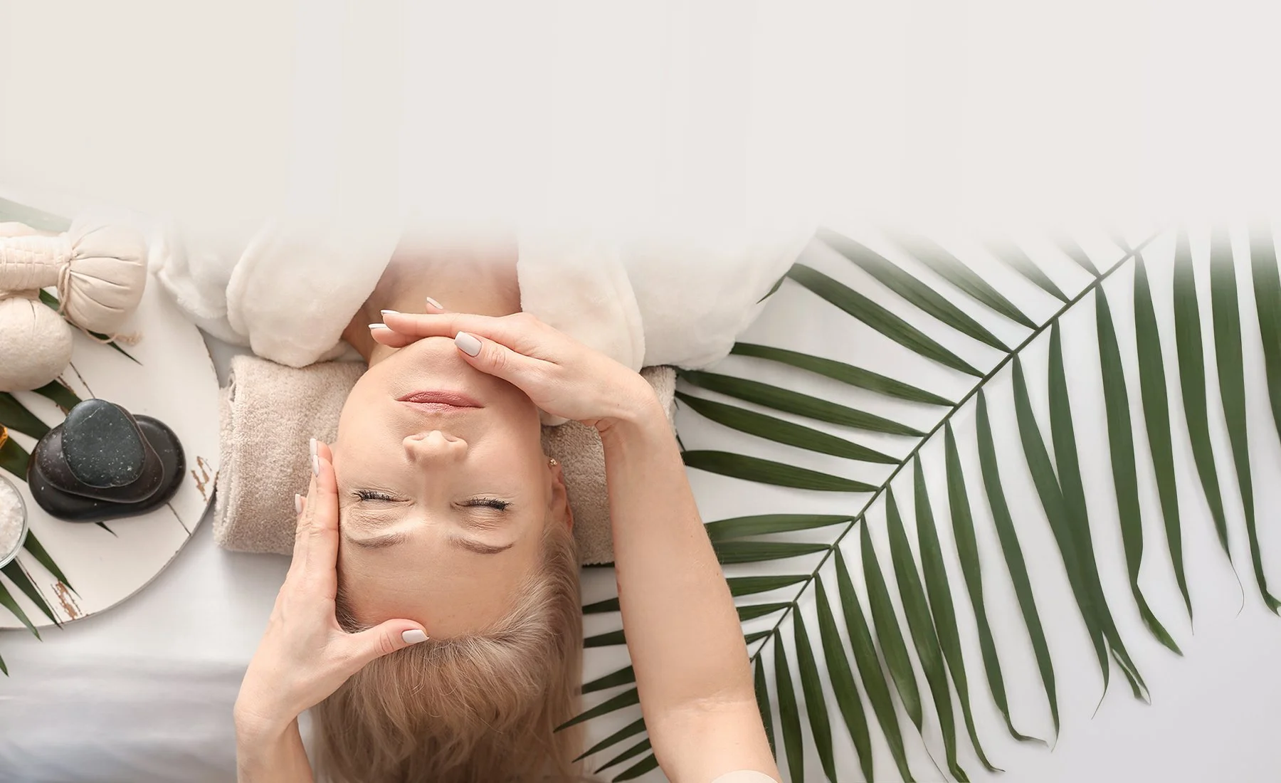 A woman lying on a massage table with her eyes closed, receiving a facial massage. There are spa stones, a massage bowl, and a towel nearby. Green palm leaves are arranged around her.