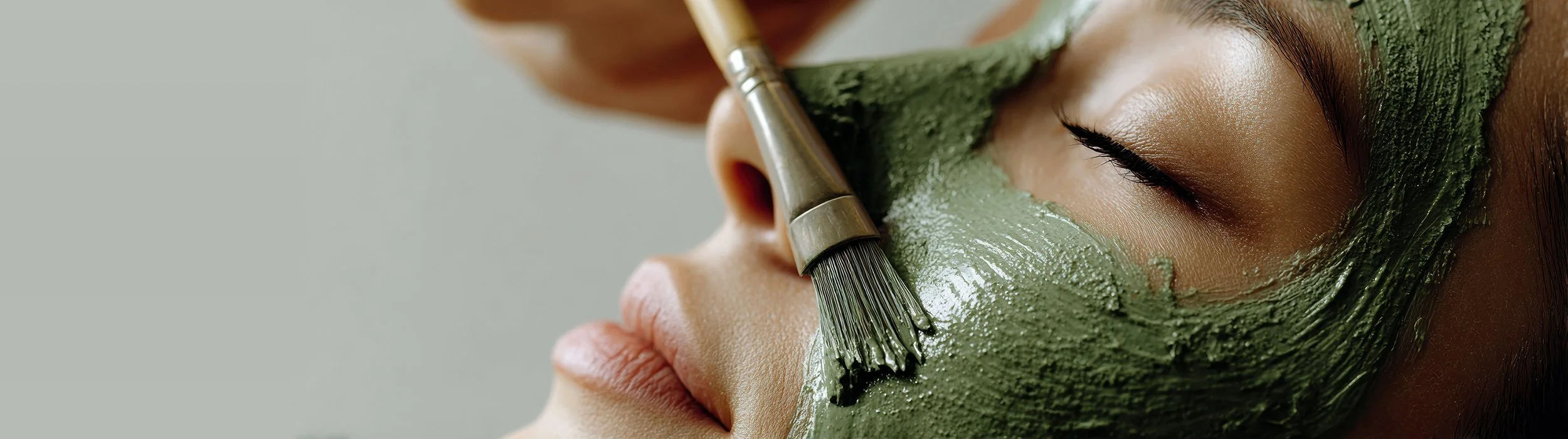 Close-up of a woman receiving a green facial mask with a brush, her eyes closed, about Renewed Life Esthetics and Head Spa.