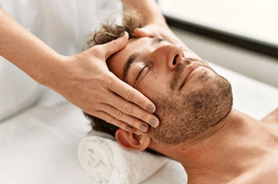 A man is lying on a massage table with eyes closed, receiving a head massage from a person whose hands are visible.