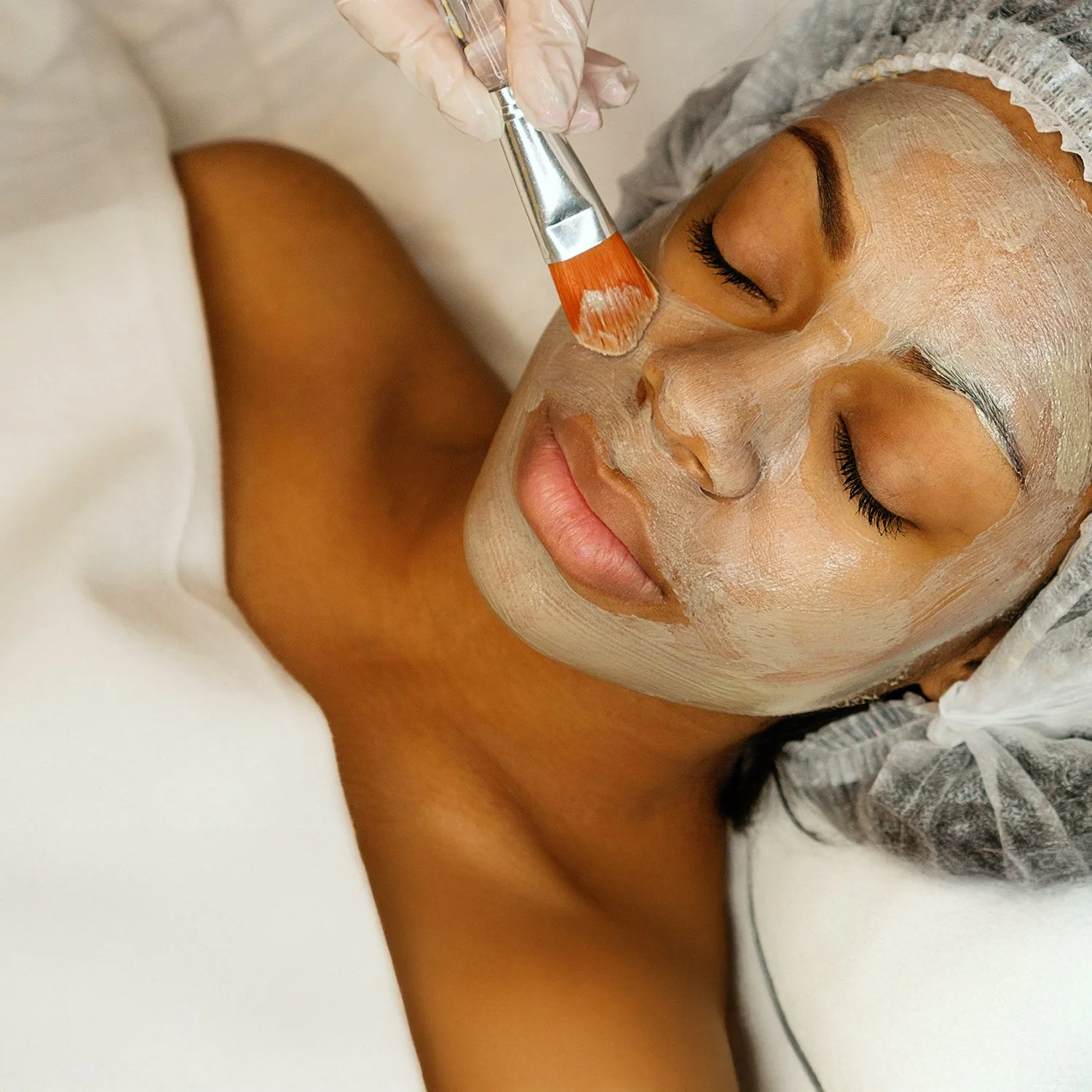 A woman receiving a facial treatment with a brush applying face mask eyes closed at Renewed Life Esthetics and Head Spa.