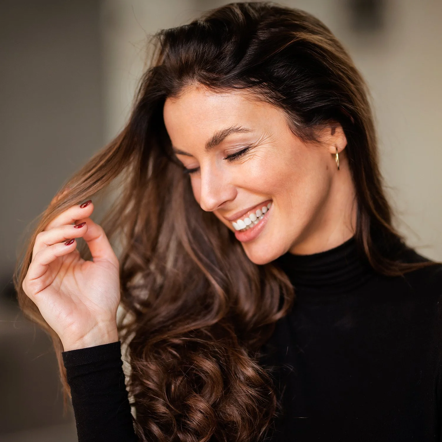 A smiling woman with long, brown, wavy hair holding her hair with hand, Hair Services at Renewed Life Esthetics and Head Spa.