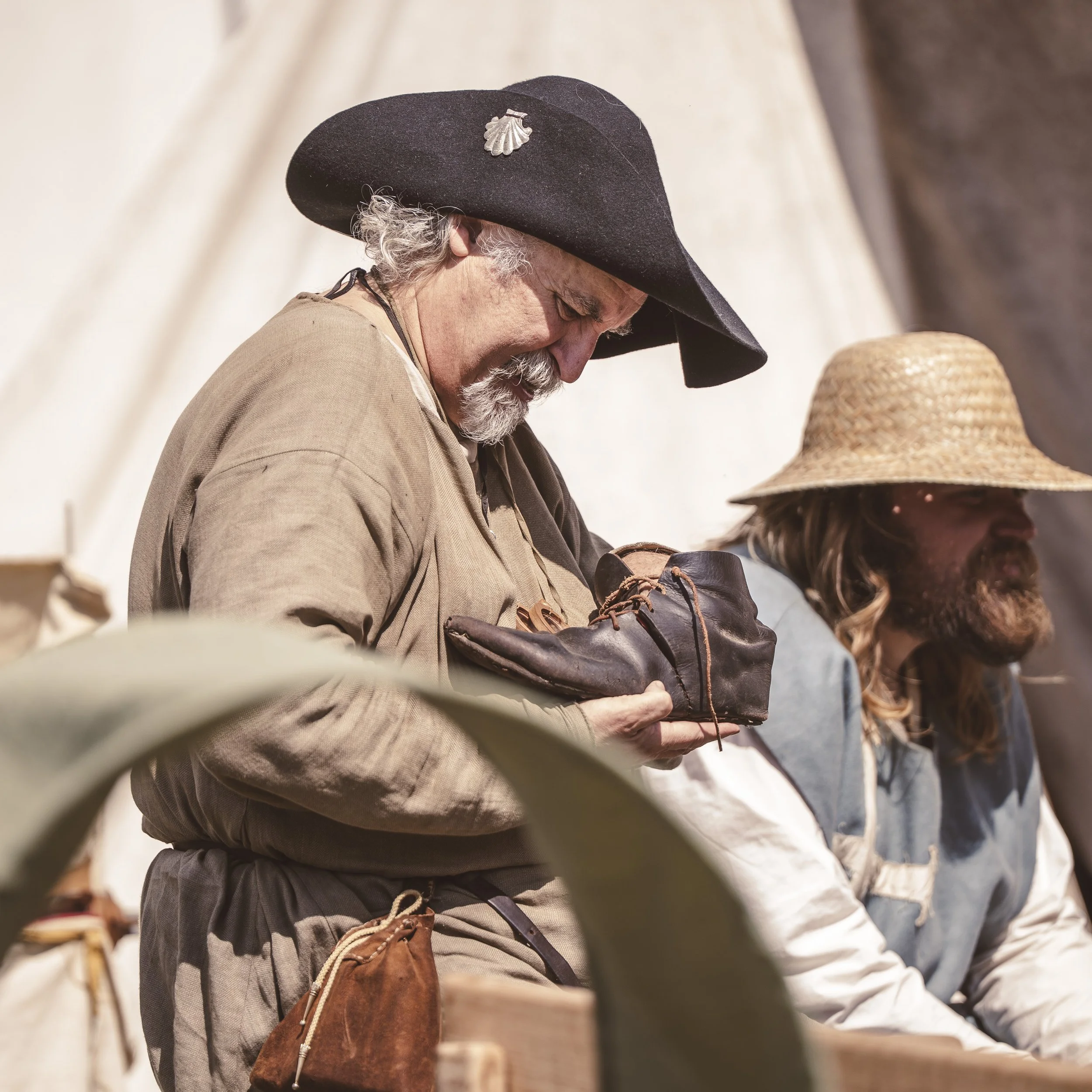 A bearded man wearing a medieval pilgrim hat and pilgrim badge inspects his leather shoe. In the background are tents and another bearded man wearing a straw hat.