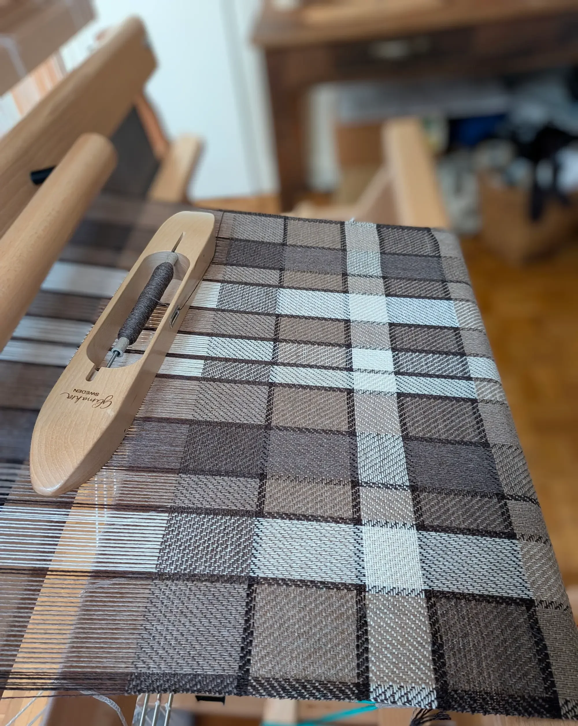 Écharpe Agate en pur cachemire en cours de tissage sur le métier, carreaux taupe, bruns et blanc cassé, navette garnie de fil brun