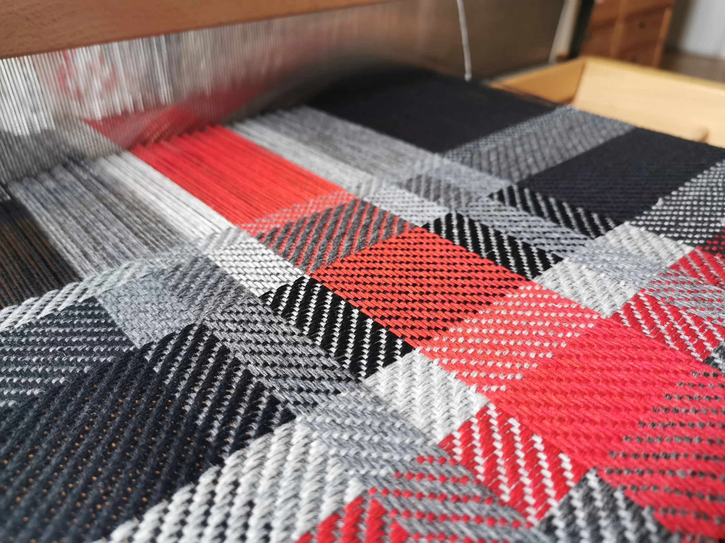 Gros plan sur l'écharpe Graphite en cours de tissage sur le métier : motif de carreaux en noir, gris, blanc et rouge vif, fils de chaîne tendus