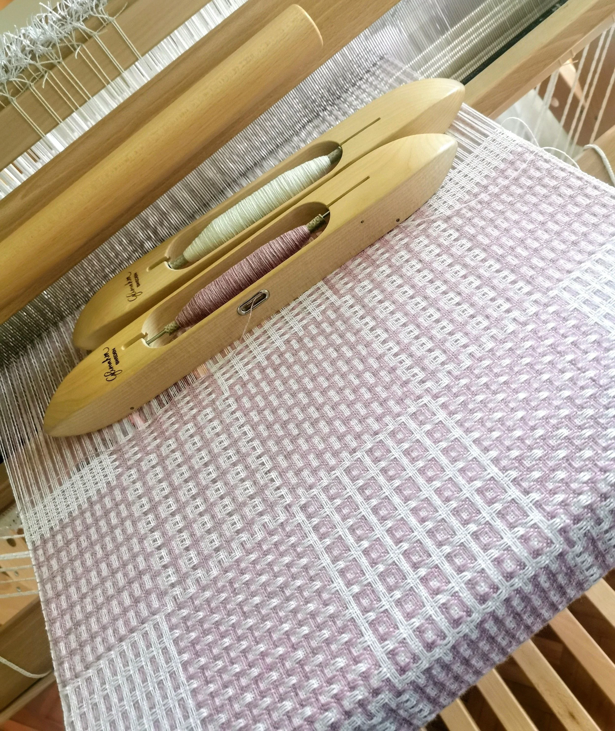 Écharpe Variation poudrée en cours de tissage sur le métier : motif à petits carrés en rose chiné et blanc, deux navettes en bois posées sur l'ouvrage