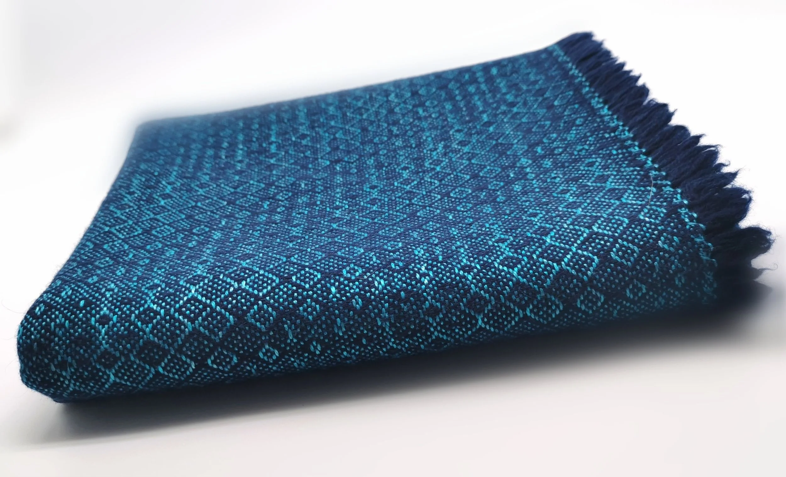 Gros plan sur l'étole Ondine en cachemire et soie pliée, motif en losanges dans un camaïeu de bleu marine et turquoise, franges visibles