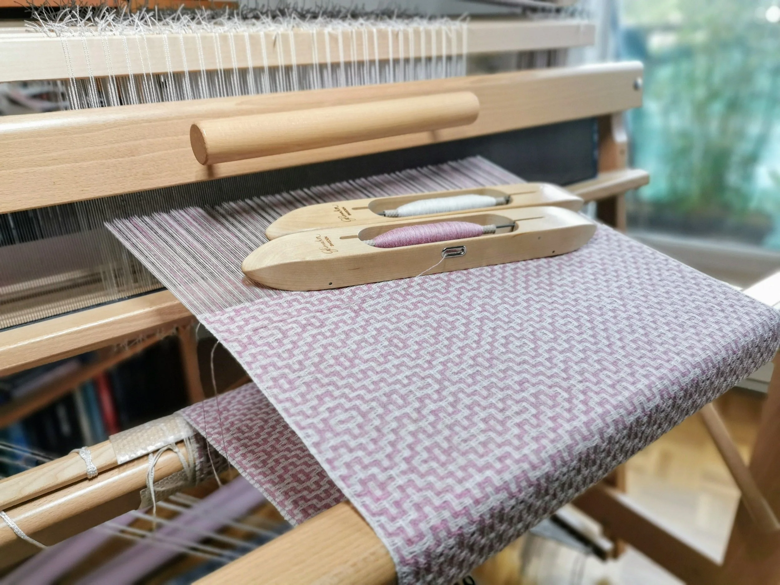 Écharpe Rosée en cours de tissage sur le métier : deux navettes en bois, l'une garnie de fil rose poudré, l'autre de fil ivoire, posées sur l'ouvrage