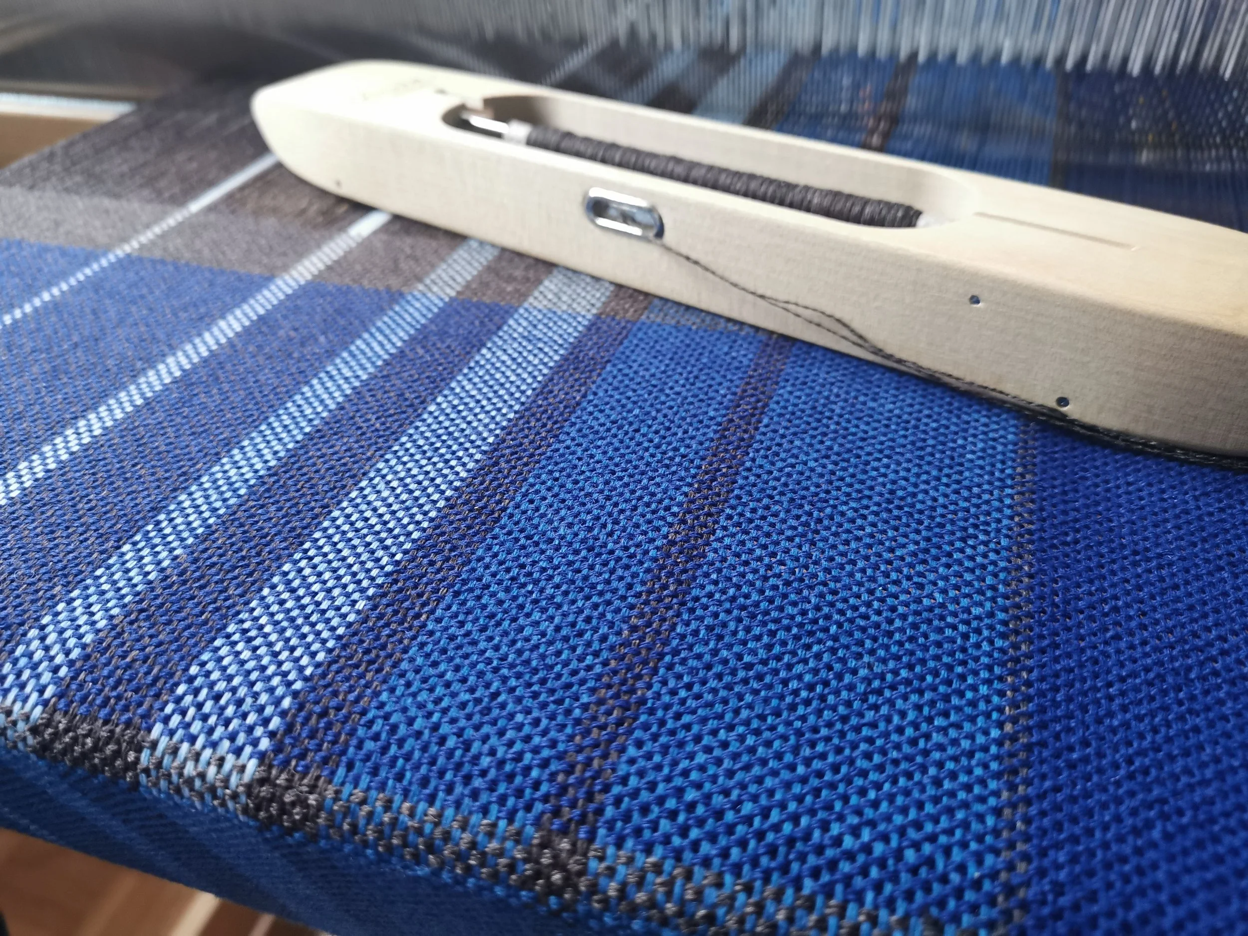 Écharpe Boréal en cours de tissage sur le métier : motif de carreaux en camaïeu de bleus, brun profond et blanc, navette en bois posée sur l'ouvrage