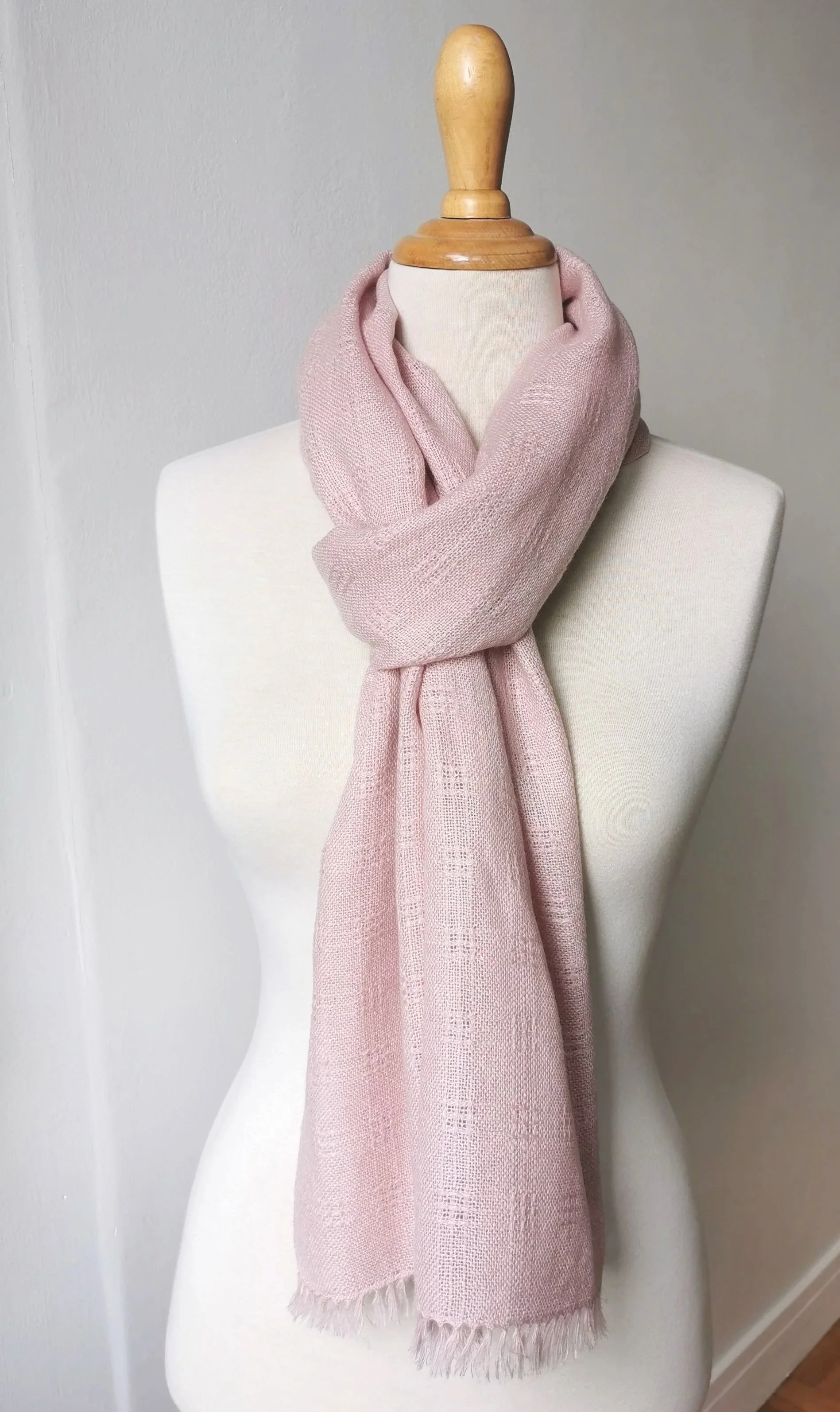 Un mannequin blanc portant une écharpe rose en laine devant un mur blanc.