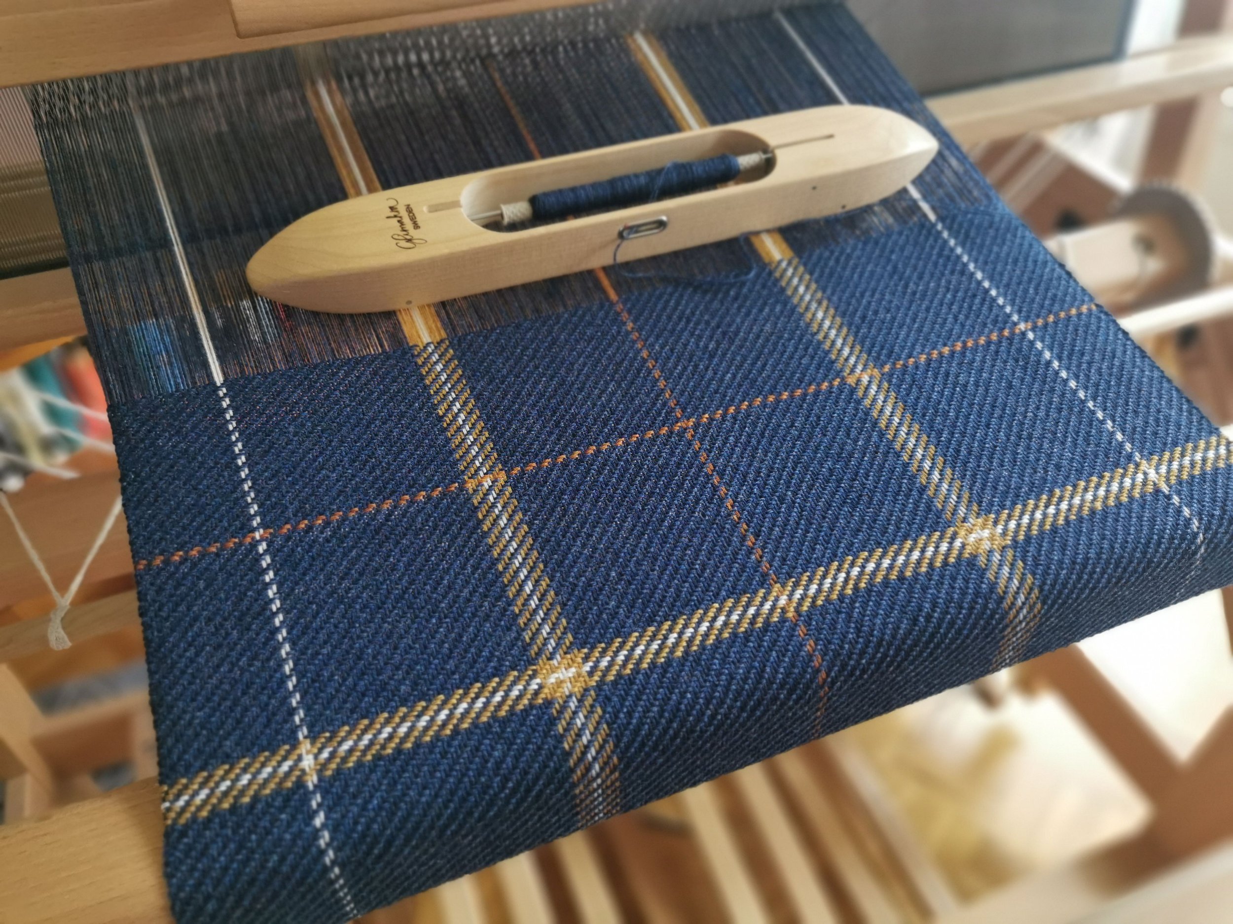 Écharpe Loch en cours de tissage sur le métier : motif de carreaux bleu profond, écru, orange et doré, navette en bois posée sur l'ouvrage