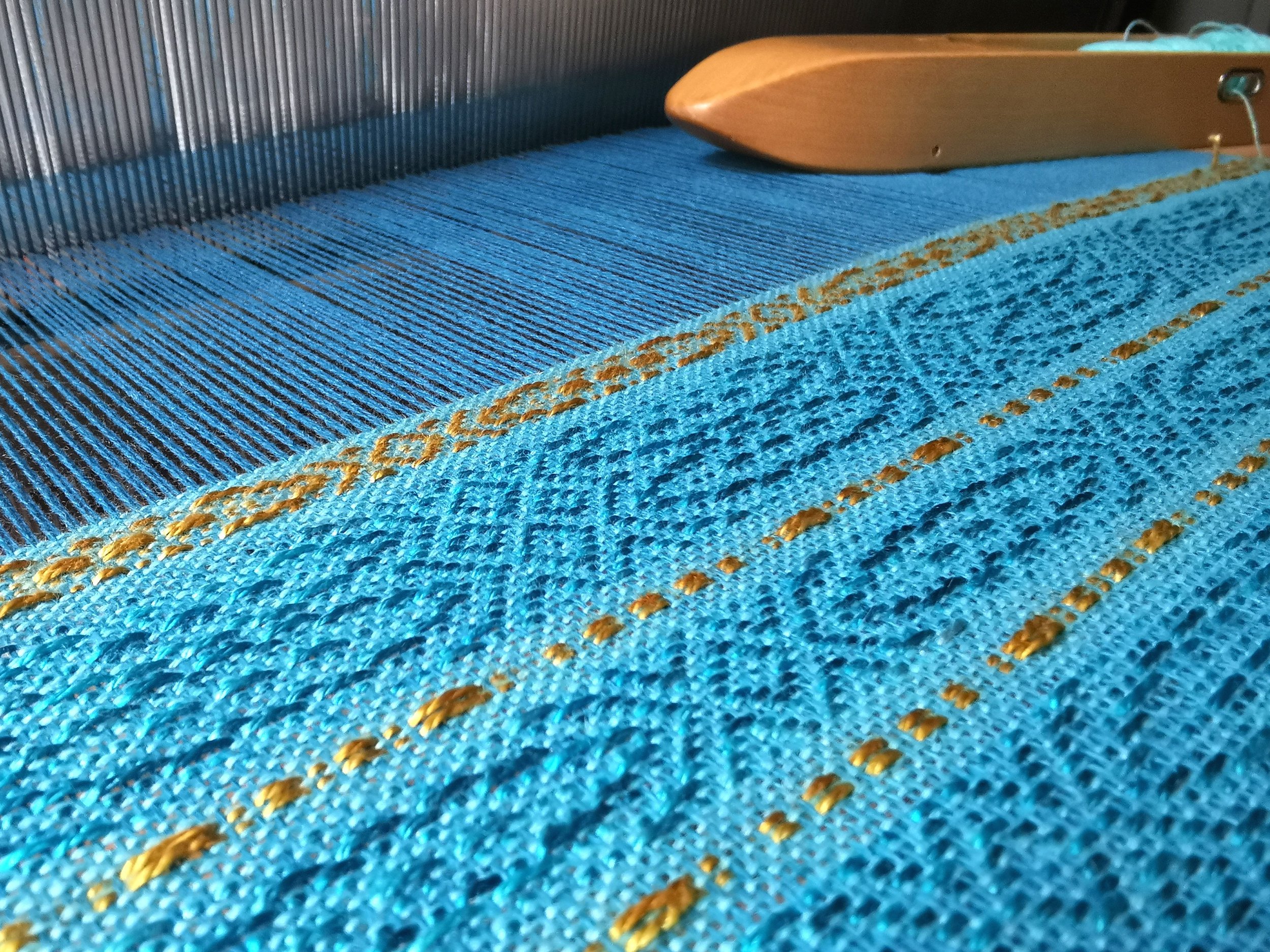 Tissu bleu avec des fils dorés, partie d'un métier à tisser avec une pièce en bois visible au sommet.